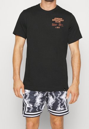 Mann trägt ein schwarzes "Jordan Breakfast Club" T-Shirt und schwarze, weiße und graue Shorts mit abstraktem Muster und steht vor einem schlichten Hintergrund.