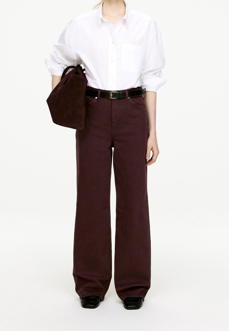 Personne portant une chemise blanche à boutons rentrée dans un pantalon large taille haute bordeaux foncé, portant un sac à main assorti en daim sur le bras.
