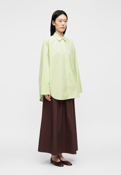 Femme debout, portant une chemise ample boutonnée vert clair, une longue jupe marron et des chaussures plates marron foncé, sur un fond blanc uni.
