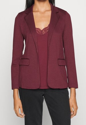 Kobieta ubrana w bordo marynarkę na cienkich ramiączkach z koronką, w czarnych spodniach, stojąca na tle jednolitego tła.