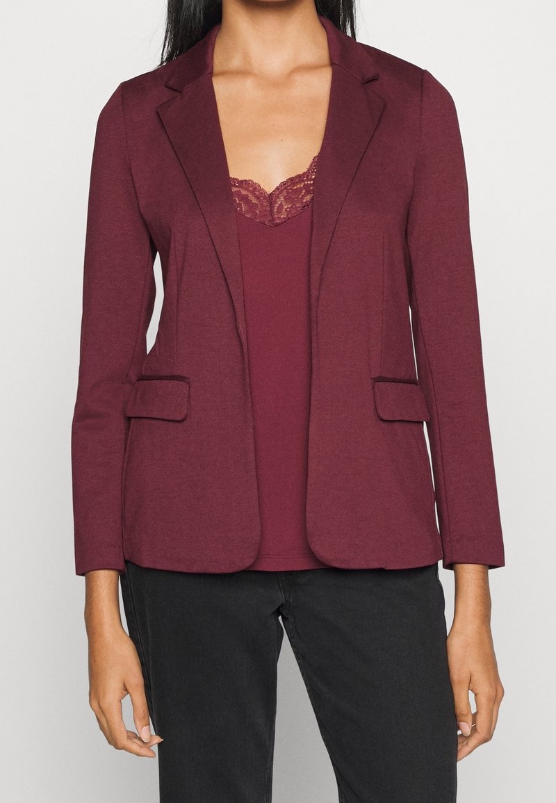 Femme portant un blazer bordeaux sur un caraco assorti à bordure en dentelle, associé à un pantalon noir, debout devant un fond uni.