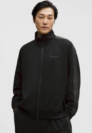 Młody mężczyzna ubrany w czarną kurtkę na suwak i spodnie z ciemnymi paskami z boku, ręce w kieszeniach, neutralne tło.