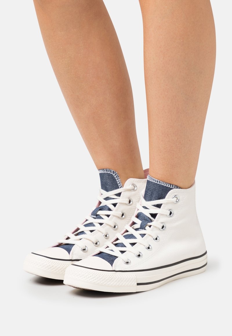 Baskets montantes blanches avec languette bleue et lacets blancs portées sur des jambes nues sur un fond clair uni.