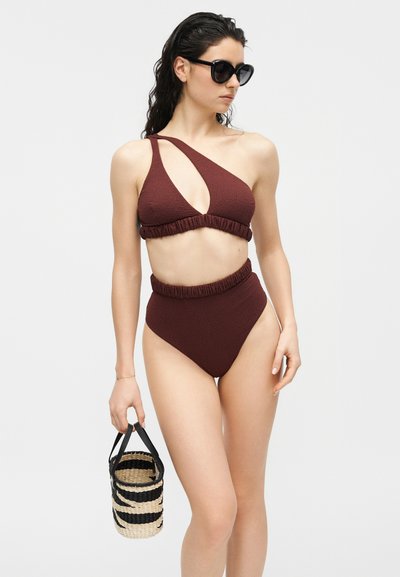 Femme en bikini bordeaux texturé à une épaule avec un bas taille haute, tenant un sac tissé à rayures noires et naturelles, portant des lunettes de soleil noires.