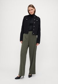 Svart denimjakke med sølvknapper, svart turtleneck og olivengrønne vide bukser, komplettert med svarte spisse sko.