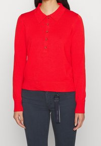 Czerwony sweter polo z długim rękawem i kołnierzykiem, z pięcioma guzikami i ściągaczami na rękawach, wykonany z gładkiego, lekkiego materiału.