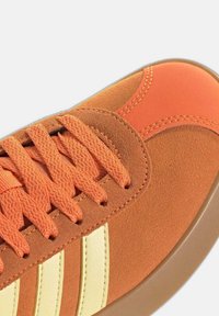 Gros plan sur une basket en daim orange avec des lacets texturés, des bandes en cuir crème et une semelle en caoutchouc sur un fond clair.