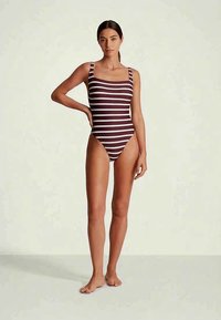 Γυναίκα που στέκεται ξυπόλητη φορώντας ολόσωμο μαγιό σε μπορντό και λευκές ρίγες, μπροστά από έναν απλό φόντο.