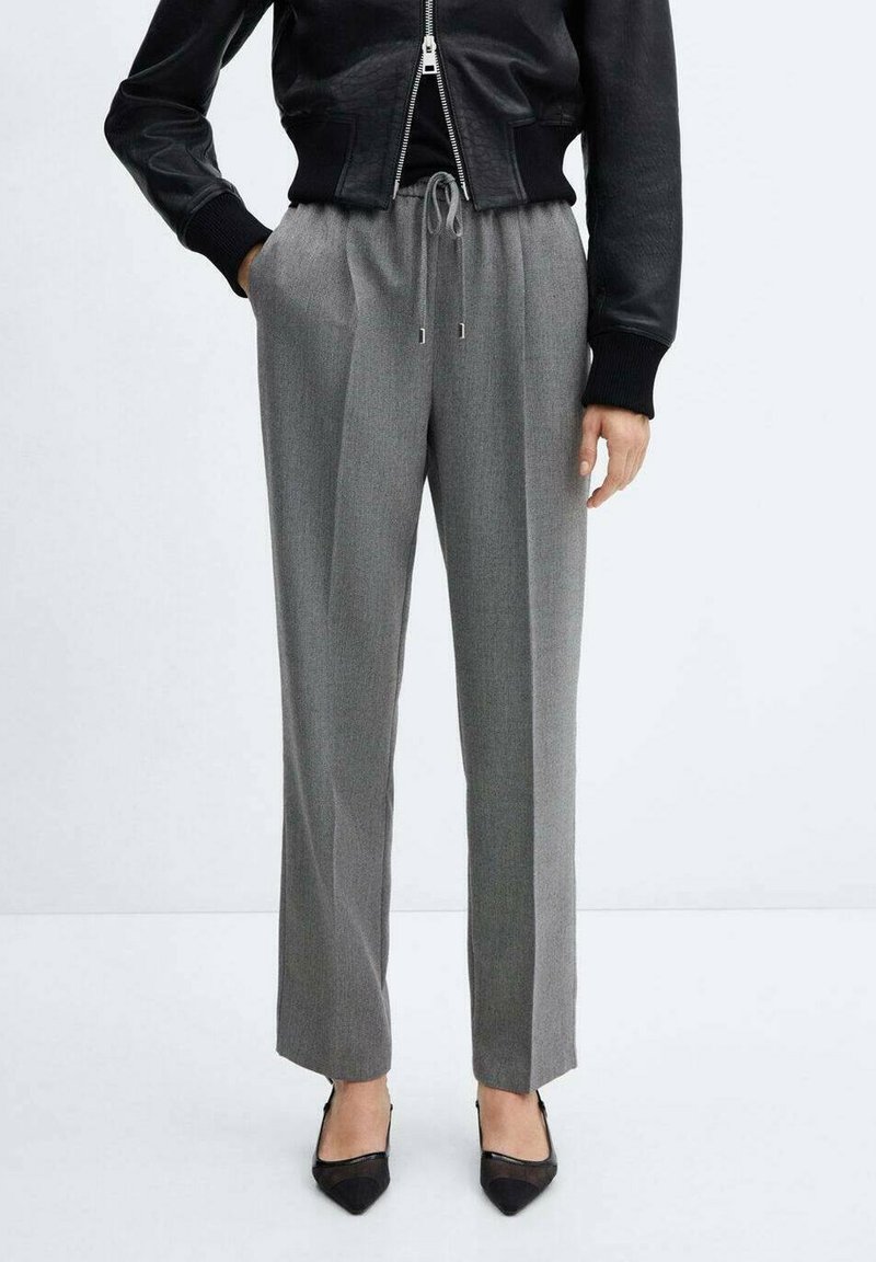 Femme portant un pantalon gris taille haute avec cordon, une veste en cuir noire zippée et des talons noirs pointus, debout sur un fond blanc.