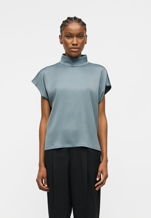 Femme aux cheveux tressés portant un haut bleu à col roulé à manches courtes amples et un pantalon large noir, debout devant un fond uni.