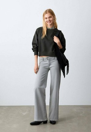 Sweat-shirt noir court, jean gris clair à jambes larges, bottines noires et sac à franges noir, sur un fond neutre.