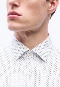 Κοντινό πλάνο στο κάτω μέρος του προσώπου και τον λαιμό ενός άνδρα που φοράει λευκό πουκάμισο με μικρό μαύρο και κόκκινο γεωμετρικό σχέδιο.