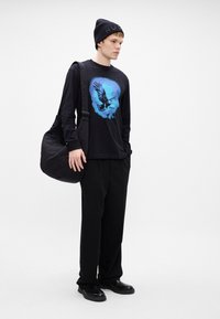 Tricou negru cu mânecă lungă, cu un grafic al unui vultur albastru, asortat cu pantaloni negri și o geantă de umăr neagră, matlasată. Completat cu o căciulă tricotată.