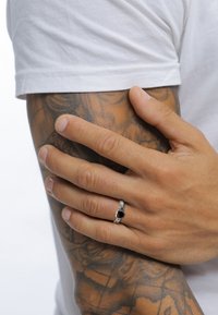 Silverring med svart sten, med en texturerad band. Handen vilar på en tatuerad arm, iförd en vit skjorta. Naturlig hudstruktur synlig.