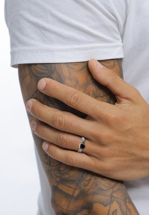 Silberner Ring mit schwarzem Stein, mit einem strukturierten Band. Hand ruht auf einem tätowierten Arm, der ein weißes Shirt trägt. Natürliche Hautstruktur sichtbar.