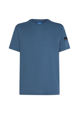 T-shirt de algodão azul com decote redondo, mangas curtas e um emblema preto na manga esquerda. Textura suave e corte descontraído.