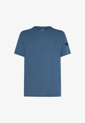 T-shirt de algodão azul com decote redondo, mangas curtas e um emblema preto na manga esquerda. Textura suave e corte descontraído.