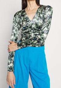 Langärmliges Oberteil mit metallischem Muster in Grün und Schwarz, das einen tiefen V-Ausschnitt und Raffungselemente aufweist. Kombiniert mit leuchtend blauen, high-waisted Hosen.