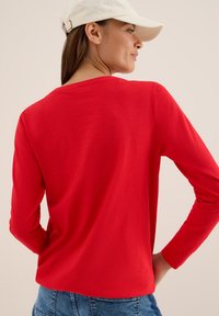 Kvinna i vit keps, röd långärmad tröja och blå jeans, står med händerna på höfterna, vänd i sidled mot en ljus bakgrund.