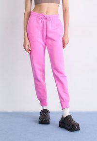Rosa joggebukser med elastisk midje og snor, prydet med en liten logo. Brukt sammen med svarte klipklapper og hvite sokker på en blå overflate.