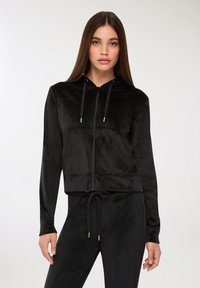 Felpa nera in velour con zip, cappuccio, tasca frontale e cordino. Presenta un design cropped e una texture liscia.