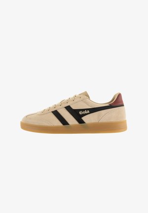 Zapatilla de ante beige con rayas negras, suela de goma y un acento en color burdeos en el talón; presenta un logo en el costado.