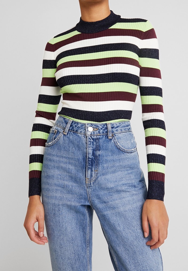 Personne portant un pull à manches longues côtelé avec des rayures horizontales en noir, blanc, vert et bordeaux, associé à un jean bleu taille haute.
