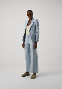 Lyseblå skreddersydd dress med en strukturert blazer og vide bukser, kombinert med en gul ribbestrikket topp og grønne slidesko.