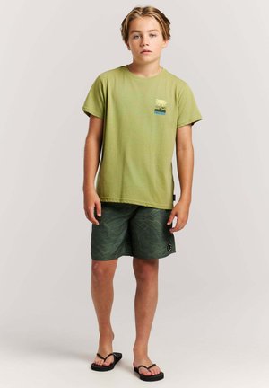 Garçon debout portant un t-shirt vert clair, un short à motifs vert foncé et des tongs noires sur un fond clair uni.