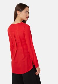 Mujer con cabello castaño liso que lleva una blusa roja de manga larga con botones dorados en el hombro y pantalones negros, de perfil.