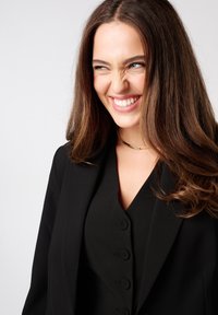 Zwarte blazer over een zwarte top met knoopdetails. Lange, golvende bruine haren; gouden choker ketting. Helderde glimlach tegen een neutrale achtergrond.