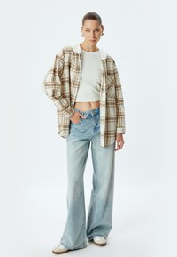 Beige und braun karierte Oversized-Hemd mit Kapuze in Weiß, kombiniert mit hellblauen weiten Jeans und beigen Slippern.