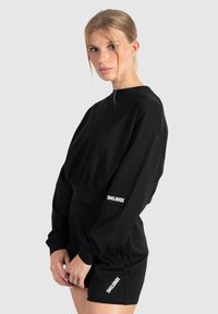 Zwart sweatshirt en shortset van zacht materiaal, met een cropped design, lange mouwen en witte "SMILODOX" logo accenten.