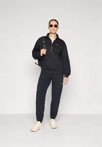 Pullover nero con zip a un quarto e logo, abbinato a pantaloni tuta neri a gamba affusolata e scarpe da ginnastica beige. Inclusi occhiali da sole e zaino.