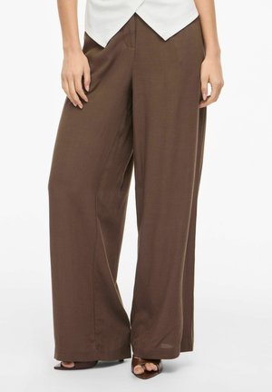 Kvinde iført bredbenede brune bukser med høj talje, kombineret med mørkebrune åbne hæle, på en hvid baggrund.
