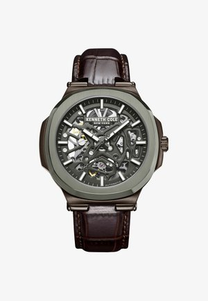 Orologio automatico con cinturino in pelle marrone, quadrante scheletrato grigio, dettagli metallici e una cassa ottagonale audace con meccanismi visibili.