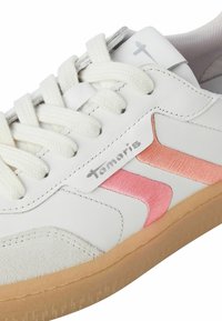Gros plan d'une basket blanche avec des lacets crème, des bandes roses brodées, une semelle en caoutchouc beige et le logo de la marque Tamari sur le côté et la languette.