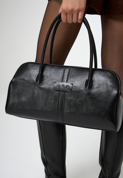 Donna che tiene in mano una borsa in pelle nera con il logo VSP, indossando collant neri trasparenti e stivali neri al ginocchio.