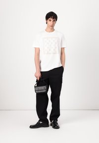 Camiseta blanca de manga corta con un intrincado patrón en beige claro, combinada con pantalones cargo negros y zapatos de cuero negros. Bolsa negra de mano.
