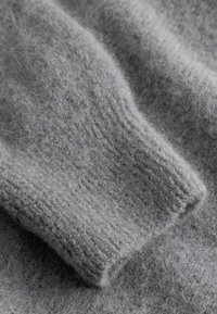 Nahaufnahme eines weichen, grauen Strickstoffs mit sichtbarer flauschiger Textur und geripptem Bündchen an einem Ärmel.