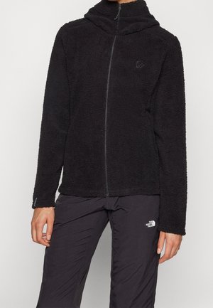 Person trägt eine schwarze Fleecejacke mit durchgehendem Reißverschluss und hohem Kragen und schwarze Outdoorhose mit The North Face Logo am Oberschenkel.