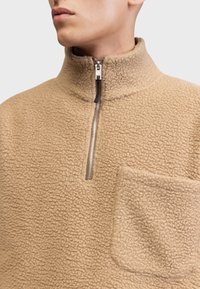 Beige fleece genser med høy krage, delvis glidelås og lomme. Strukturert overflate med et mykt utseende.