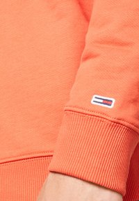 Felpa arancione con tessuto a costine e polsini a coste. Presenta una piccola etichetta con logo rosso, bianco e blu sulla manica sinistra.
