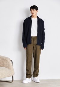 Cardigan côtelé bleu marine, chemise blanche à col ras du cou, pantalon olive à cordon de serrage et baskets blanches, sur un fond neutre avec une chaise texturée.