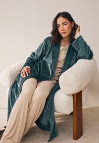 Zamatový tyrkysový župan voľne prehodený cez osobu oblečenú v béžový rebrovaný outfit, sediacu na nadýchanej bielej stoličke s drevenými nohami.