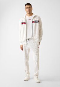 Weißer Trainingsanzug aus strukturiertem Stoff mit Reißverschlussjacke, roten und schwarzen Streifen, Logo auf der Brust und passenden Hosen mit Seitentaschen.