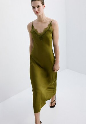 Femme portant une robe nuisette en satin vert olive avec bordure en dentelle et des sandales noires à bout carré, debout dans une pièce blanche minimaliste.