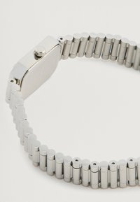 Ασημένιο ρολόι από ανοξείδωτο ατσάλι με ορθογώνιο καντράν και μεταλλικό μπρασελέ που διαθέτει στρογγυλεμένους συνδέσμους και κορώνα στη πλευρά.