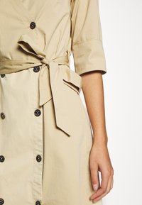 Beiges Kleid mit Wickeldesign, kurzen Ärmeln und schwarzen Knöpfen. Mit einem gebundenen Gürtel in der Taille und einer Seitentasche. Glatte Stofftextur.