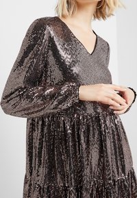 Robe scintillante en sequins bronzés avec un décolleté en V, des manches longues et un design à volants en étages. Agrémentée de poignets en forme de vagues.